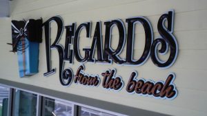 “Regards from the Beach” dimensional storefront letters with a decorative bow graphic mounted on a light-colored facade.