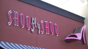 Pink “Shoo Fly Shoo” storefront lettering with a large high-heel graphic mounted on the building facade.