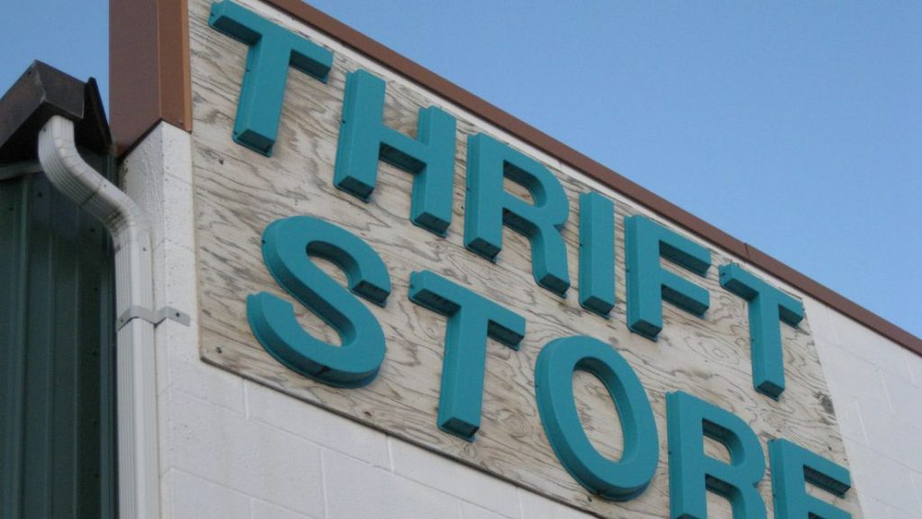 A slightly low-angle shot of a "THRIFT STORE" sign mounted on the exterior of a building. The sign features raised, teal-colored 3D letters mounted on a lighter, possibly wooden or textured, rectangular panel.