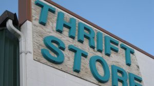 A slightly low-angle shot of a "THRIFT STORE" sign mounted on the exterior of a building. The sign features raised, teal-colored 3D letters mounted on a lighter, possibly wooden or textured, rectangular panel.