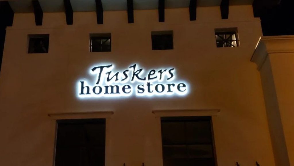 Illuminated Tuskers Home Store lettering glowing against the building facade at night.