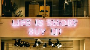 A bright pink neon sign in a store window reads "LIFE IS SHORT BUY IT!", with track lighting below it illuminating a white tiled wall.