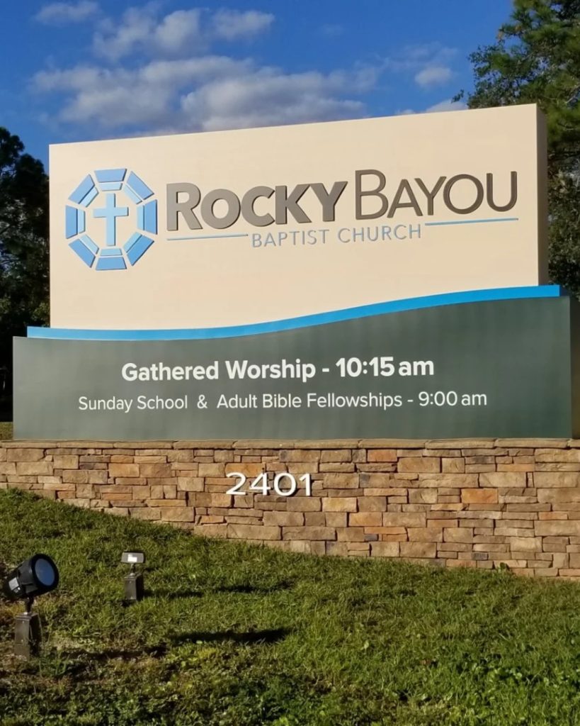 Custom church monument sign design featuring the "Rocky Bayou Baptist Church" name and service times on a modern stone base with a curved blue accent. This professionally crafted monument sign blends stone and acrylic for high visibility and architectural appeal.