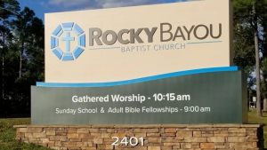Brick monument sign for Rocky Bayou Baptist Church, displaying service times and featuring a modern design with a stone base and blue accents.
