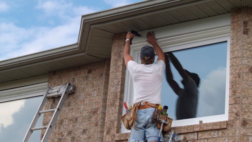 A worker installing a metal awning above a residential window, using tools and a ladder against a brick wall.