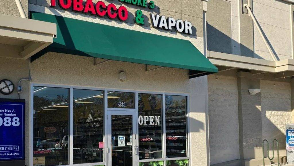 Exterior storefront of a smoke shop featuring green awning signs for business in Land O' Lakes FL, with bold red and white lettering for “Tobacco & Vapor” above the entrance.
