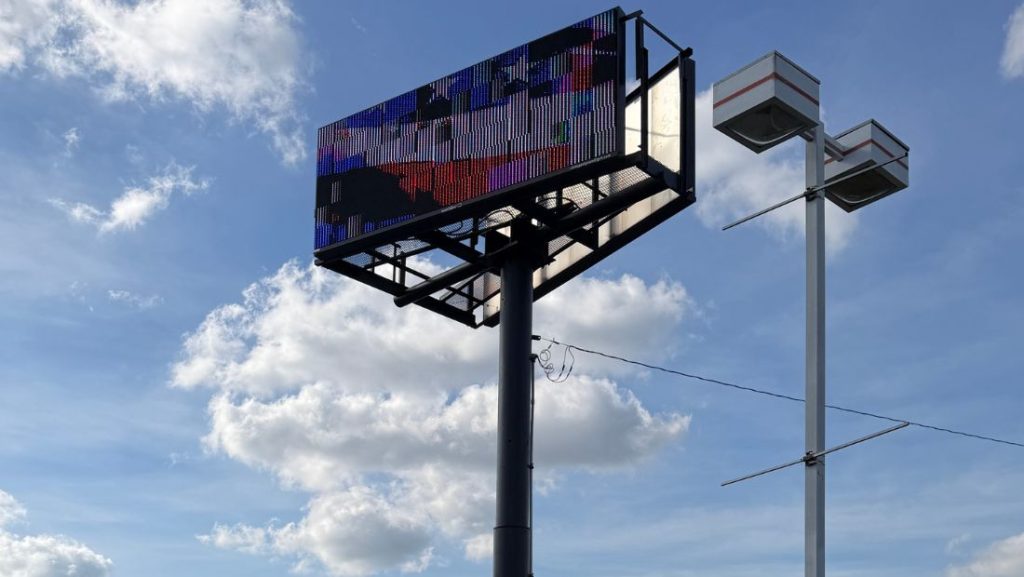 An outdoor LED billboard mounted on a tall pole against a bright blue sky with clouds, showcasing a digital display, often installed by businesses to boost visibility and advertising reach.