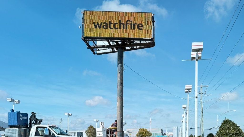 A technician working on a large Watchfire LED digital billboard, with the bright yellow "watchfire" logo displayed on the screen against a clear blue sky.
