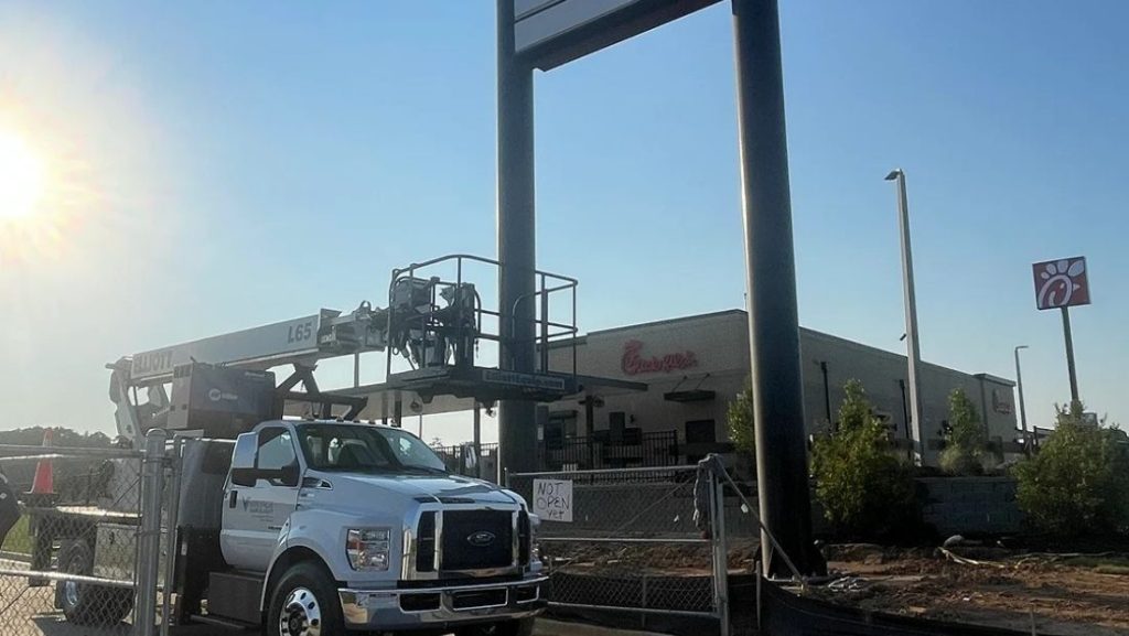 Work truck and lift installing a large commercial sign in front of a Chick-fil-A building on a sunny day.
