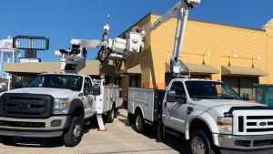 Two white service trucks with bucket lifts parked outside a commercial building as reliable installers for hurricane resistant signage in coastal areas perform maintenance on exterior signs under clear blue skies.
