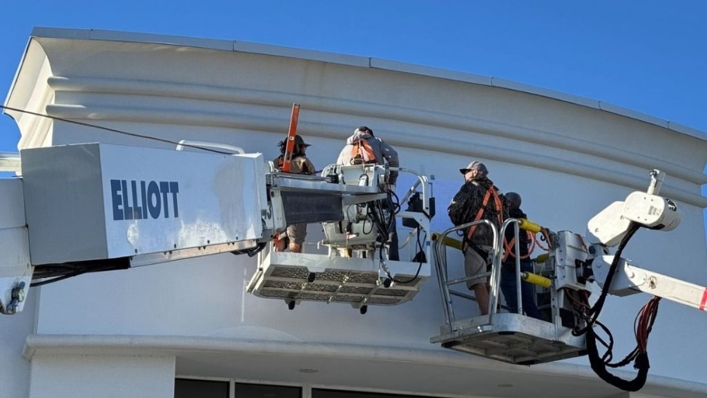 Crew of workers in safety harnesses installing signage on a building façade using Elliott boom lifts under a clear blue sky.