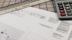 Close-up of sign construction plans on a table with a scale ruler and calculator, showing technical drawings, notes, and permit details.