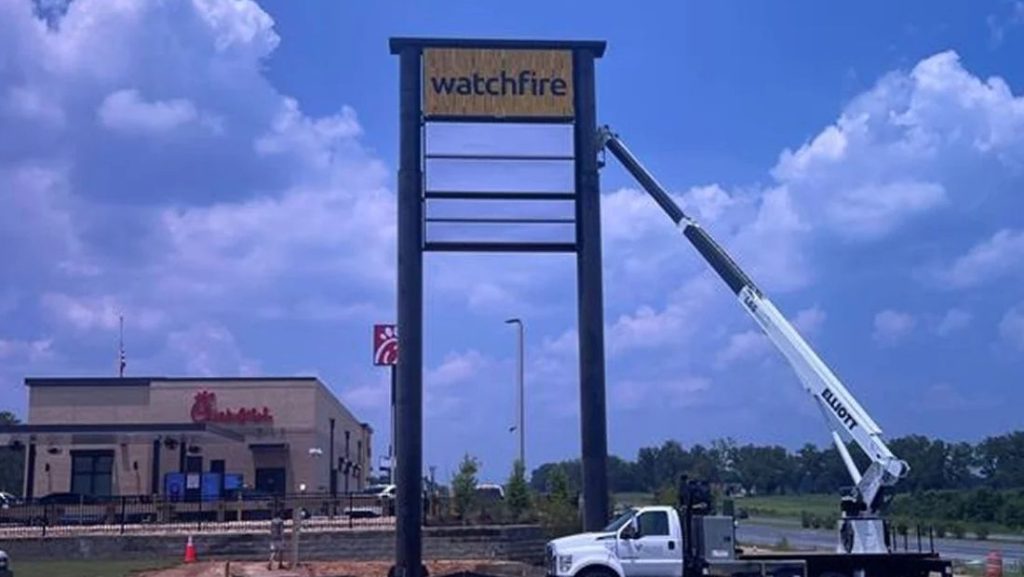 Watchfire digital signs being installed with a crane in front of a Chick-fil-A location. The sign is mounted on a tall structure, with the Watchfire logo visible.