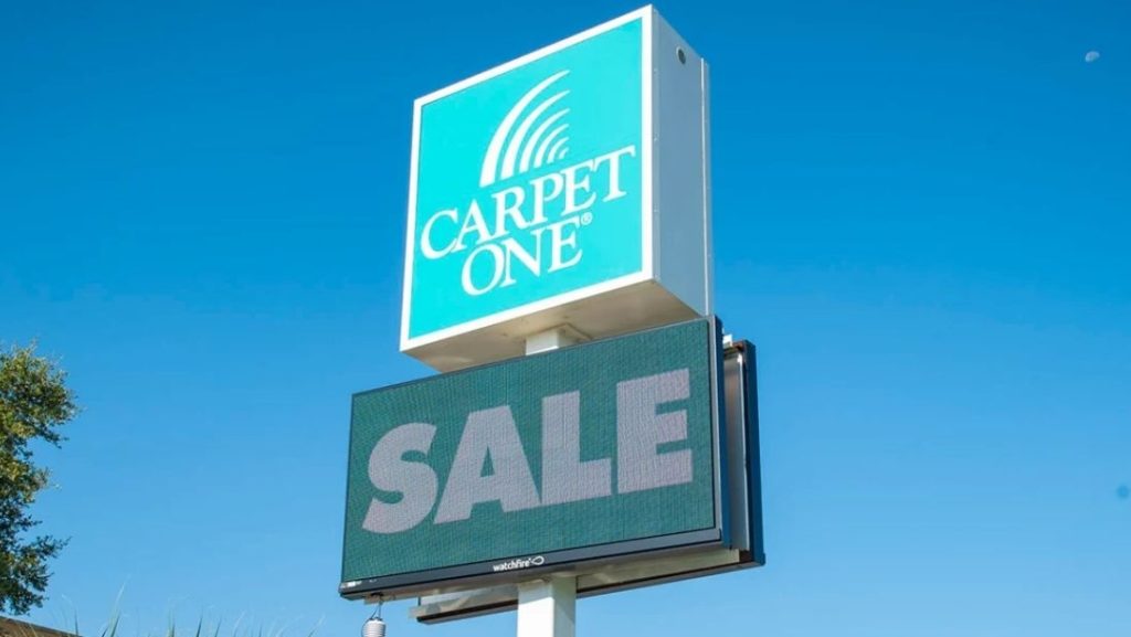 Retail digital signage display showing the word 'SALE' in large bold letters on a digital screen below the Carpet One store logo.