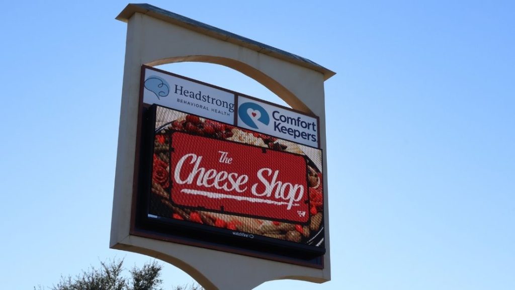 Digital signage for restaurants showcasing 'The Cheese Shop' with an eye-catching display on the digital screen, surrounded by branding from Headstrong Behavioral Health and Comfort Keepers.