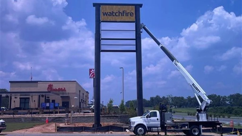 Retail digital signage installation in progress, showing a worker using a crane to mount a Watchfire digital display above a store, with a Chick-fil-A restaurant in the background.