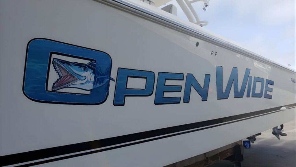 Custom boat lettering featuring the “Open Wide” name and shark logo in blue vinyl graphics on the side of a white fishing boat hull.