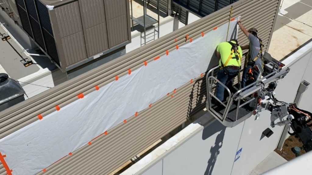 Technicians in a lift basket using professional sign installation tools to mount a large commercial sign panel on an exterior building wall.