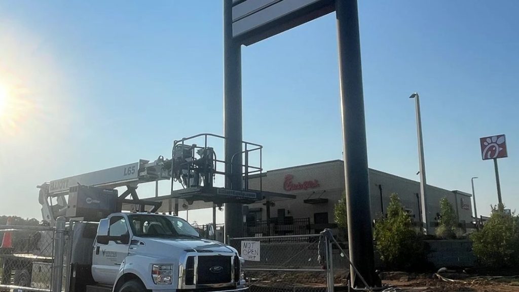 Bucket truck and crew installing a large elevated sign structure near a commercial building, preparing a tall pylon sign for high visibility.