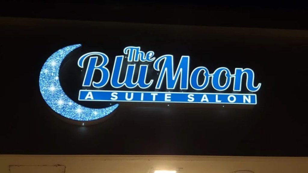Illuminated storefront sign for The Blu Moon Suite Salon with blue channel letters and crescent moon graphic on a dark background, showing how to install channel letters for night visibility signage.