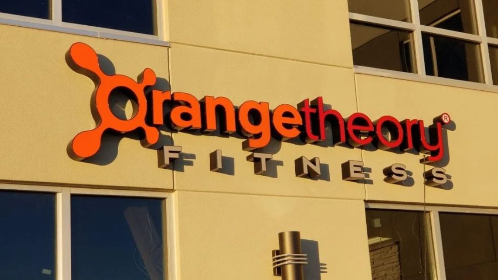 Exterior storefront sign for Orangetheory Fitness featuring illuminated orange channel letters mounted on a beige building, example of how to install channel letters for commercial signage.