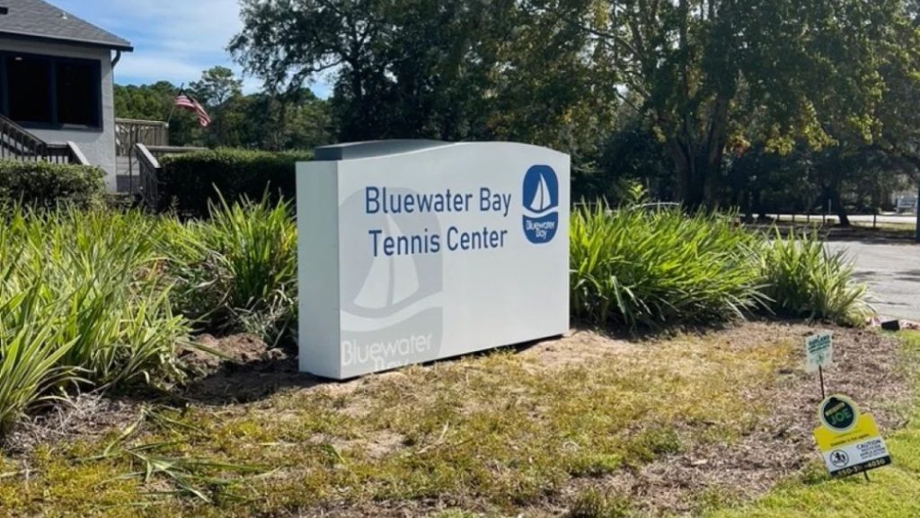 Standard daytime view of a small monument sign reading 'BLUEWATER BAY TENNIS CENTER', featuring a clean stone base and surrounding plants