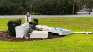 Damaged monument sign lying on grass beside a broken brick base, showing an outdoor sign structure needing repair or replacement.