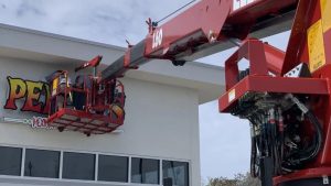 Technician installing a storefront sign using a boom lift on a commercial building exterior.