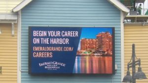 Outdoor LED display showing a hiring message on a commercial building.